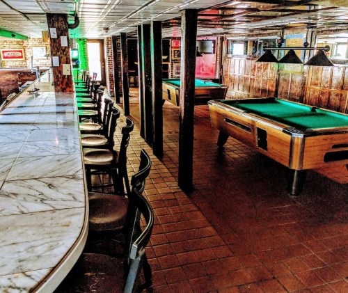 A spacious bar with a marble counter, empty stools, and pool tables in a cozy, well-lit setting.