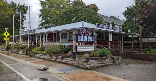 A quaint general store with a blue and red sign, surrounded by greenery on a rainy day.