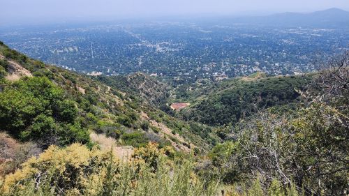 A panoramic view of a lush green valley and distant cityscape from a mountain trail.
