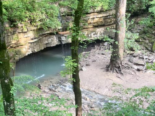 A serene natural scene featuring a rocky overhang, flowing water, and lush green trees surrounding a small creek.