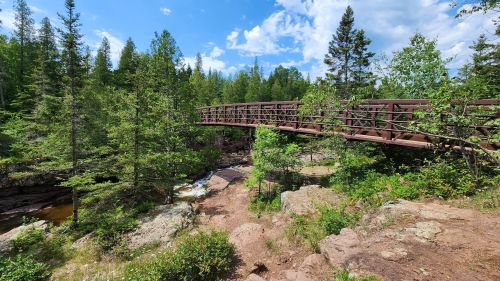 A wooden bridge spans a rocky area surrounded by lush green trees and a clear blue sky.