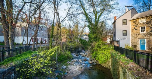A serene stream flows through a lush green area, flanked by historic buildings and trees in early spring.