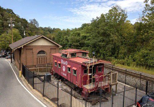 A red B&O caboose parked next to a brick building, surrounded by a fence and trees in the background.
