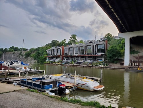 View of a river with boats docked, modern townhouses along the shore, and a bridge overhead under a cloudy sky.