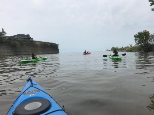 Three kayakers paddle on calm water near a rocky shoreline under a cloudy sky.