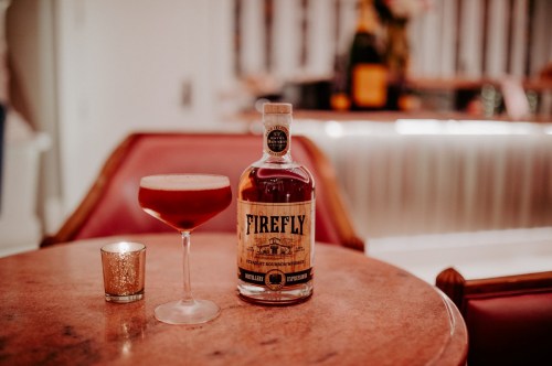 A bottle of Firefly whiskey next to a cocktail in a glass, on a round table with a candle in a holder.