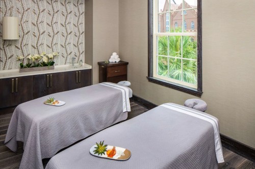 A serene spa room with two massage tables, decorated with fresh flowers and a view of palm trees outside.