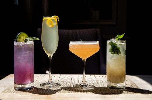 Four colorful cocktails on a table: a purple drink, a yellow drink with lemon, a peach cocktail, and a green mint drink.