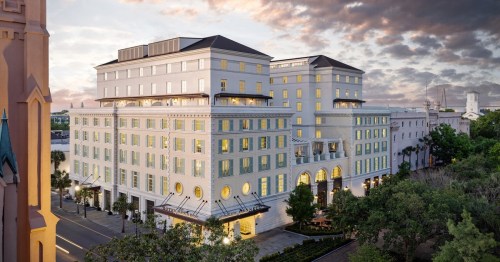 A modern hotel with large windows and a light-colored facade, surrounded by greenery under a cloudy sky.