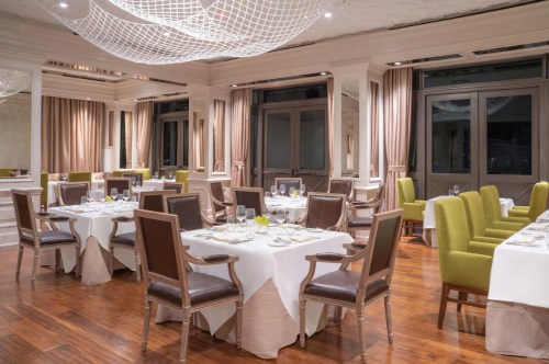 Elegant restaurant interior with wooden floors, white tablecloths, and green and brown chairs under a decorative light fixture.