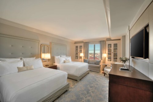 A spacious hotel room with two beds, a seating area, and large windows offering a view. Elegant decor and soft lighting.