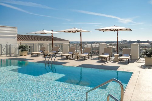 Rooftop pool with lounge chairs and umbrellas, overlooking a city skyline under a clear blue sky.