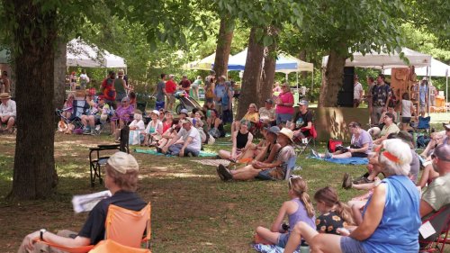 A crowd of people sitting on the grass under trees, enjoying an outdoor event with tents in the background.