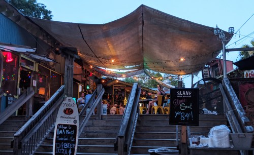 A lively outdoor restaurant with colorful lights, a large canopy, and wooden stairs leading to a bustling dining area.