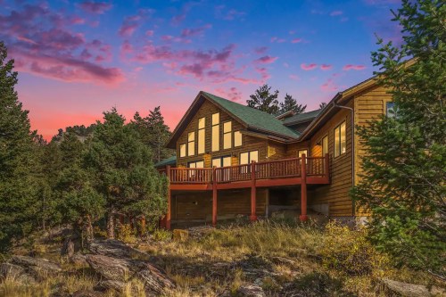A wooden house with large windows, surrounded by trees, against a colorful sunset sky.