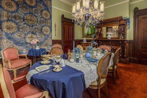 Elegant dining room set with a blue tablecloth, fine china, and a chandelier, featuring ornate decor and floral arrangements.