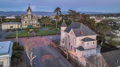 Aerial view of a historic Victorian-style building and a pink house, surrounded by trees and mountains in the background.
