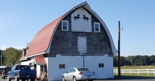 A red-roofed barn with horse silhouettes, surrounded by parked cars and open fields under a clear blue sky.