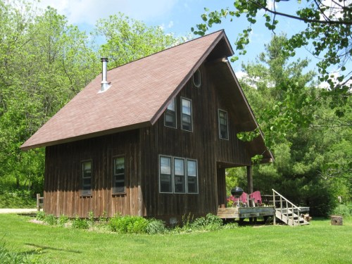 A rustic wooden house with a sloped roof, surrounded by greenery and a small deck with chairs.
