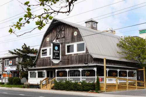 A rustic wooden building with a peaked roof, featuring a sign for "Barn 34" and surrounded by greenery.