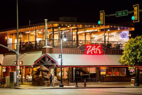 A lively restaurant at night, featuring outdoor seating and bright neon signs, located at a street intersection.