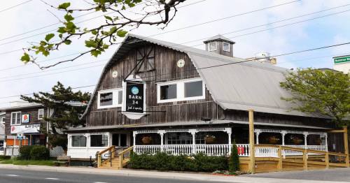 A rustic wooden building with a peaked roof, featuring a sign that reads "Barn 34" and surrounded by greenery.