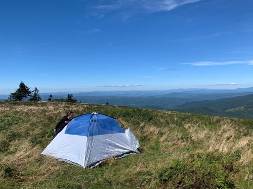 A person sets up a blue and gray tent on a grassy hilltop with a panoramic view of distant mountains and a clear sky.