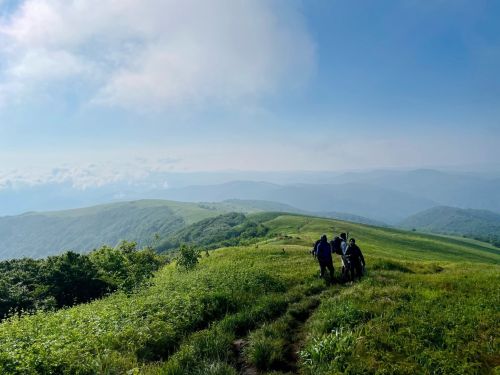 Two hikers walking along a grassy trail with rolling hills and a blue sky in the background.