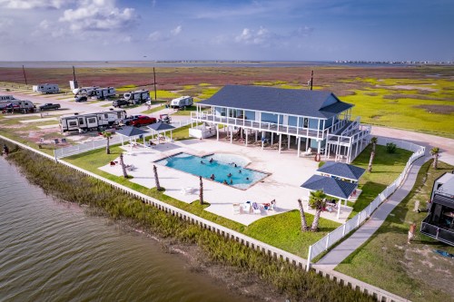 Aerial view of a coastal house with a pool, surrounded by palm trees and RVs, near a marshy area and blue sky.