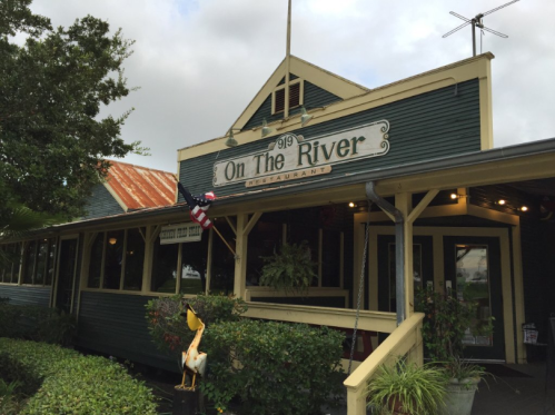 Exterior of "On The River" restaurant, featuring a green facade, porch, and American flag. Lush greenery surrounds the entrance.