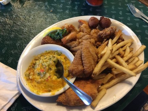 A large plate of fried seafood, including shrimp, fish, hushpuppies, and fries, served with a side of dipping sauce.