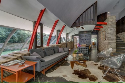 Modern living room with a gray sofa, glass coffee table, and unique architecture featuring red beams and large windows.
