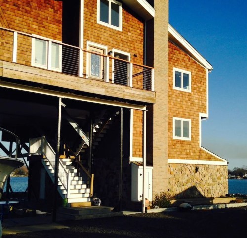 A modern two-story house with wooden siding, a balcony, and a staircase leading to the entrance, by a body of water.