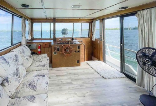 Cozy boat interior with a steering wheel, sofa, and large windows overlooking the water.