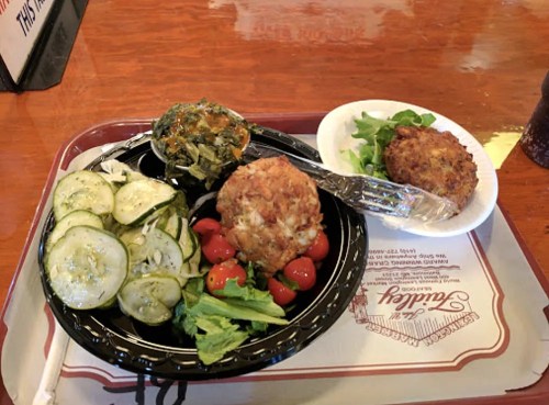 A tray with a serving of vegetables, a crab cake, and a side salad, featuring sliced cucumbers and cherry tomatoes.
