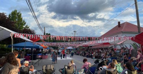 A lively street scene with people sitting under tents, enjoying a community event decorated with flags and bunting.