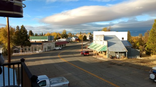A quiet small town street with a few buildings, trees, and a cloudy sky in the background.