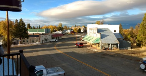A quiet small town street with shops, trees in autumn colors, and a cloudy sky in the background.