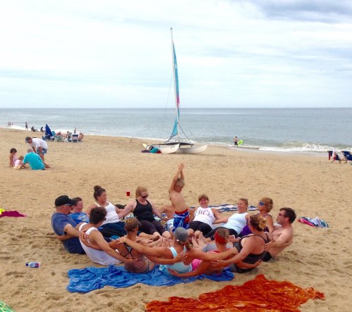 A group of people sitting in a circle on the beach, with a sailboat in the background and others enjoying the shore.