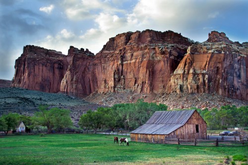 A rustic barn in a green field with horses, set against towering red rock cliffs and a cloudy sky.