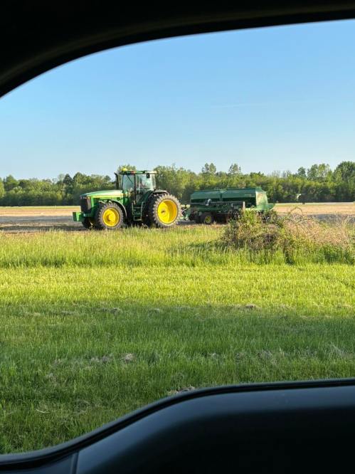 A green tractor pulling a trailer in a field, with trees and blue sky in the background.