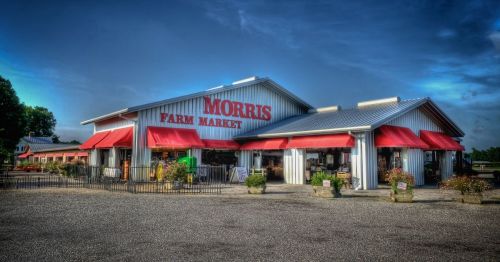 A large farm market building with red awnings and signage, surrounded by a gravel lot and greenery.