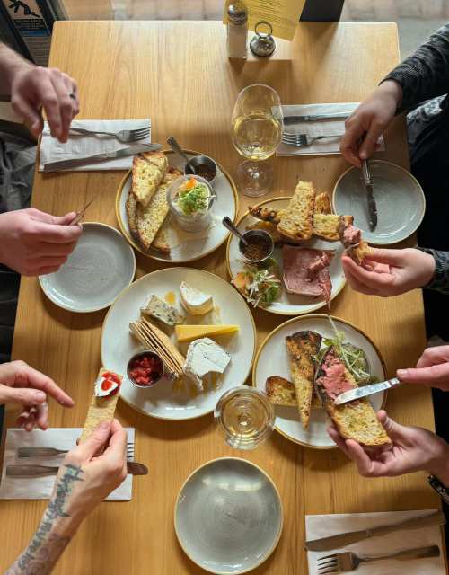 A table with various dishes, including bread, cheeses, meats, and condiments, as hands reach for the food.