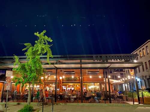A lively restaurant at night, with outdoor seating and warm lights illuminating the brick facade and nearby tree.