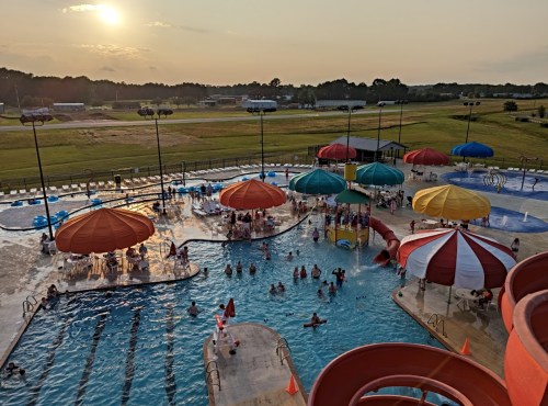 Aerial view of a water park with colorful umbrellas, pools, and people enjoying the sunset in the background.