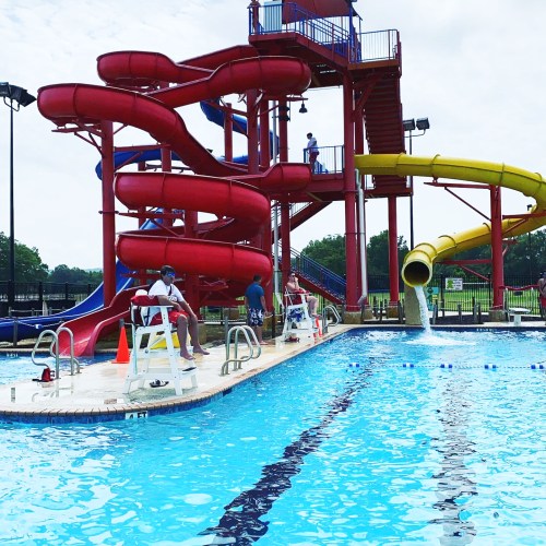 A colorful water slide towers over a swimming pool, with people enjoying the area on a sunny day. Lifeguards are present.