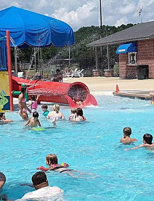 Children playing in a water park pool with a slide and splash features under a blue sky.