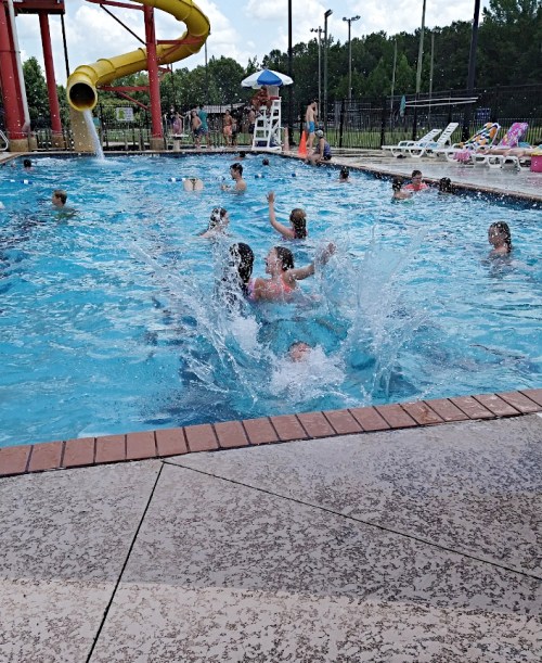 A lively pool scene with children splashing and playing, a water slide in the background, and sunbathers nearby.