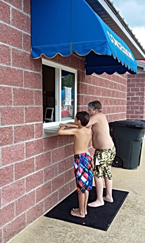 Two boys in swim trunks stand at a concession window, eagerly waiting for their order. A trash bin is nearby.