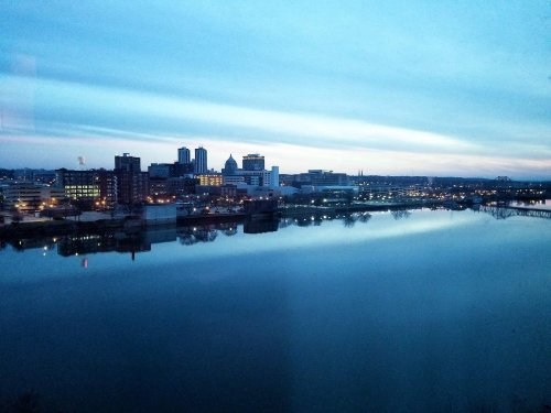 City skyline at dusk reflected in a calm river, with buildings and lights creating a serene atmosphere.